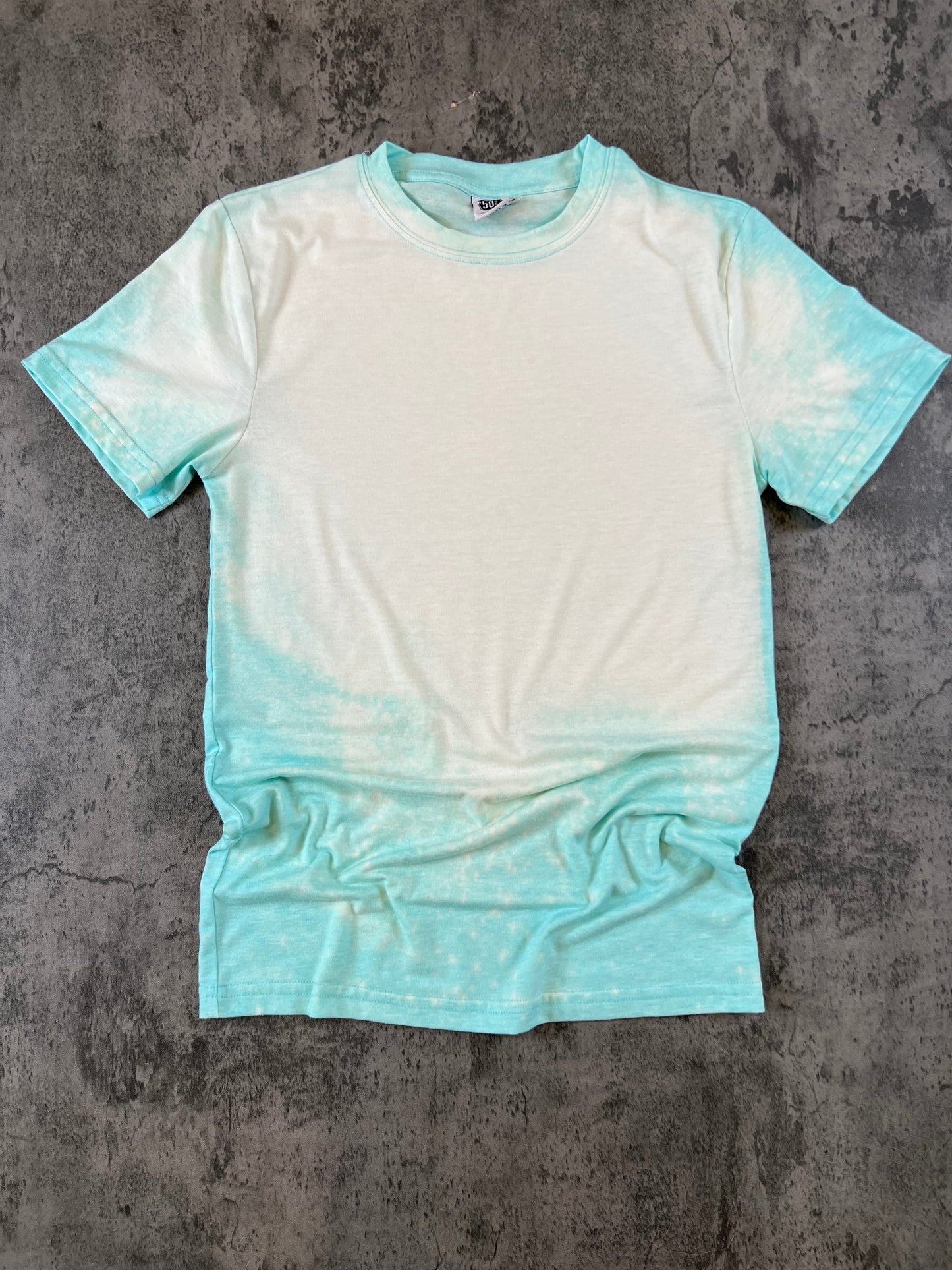 Aqua blue t-shirt with bleached front chest area, laying flat on a concrete background
